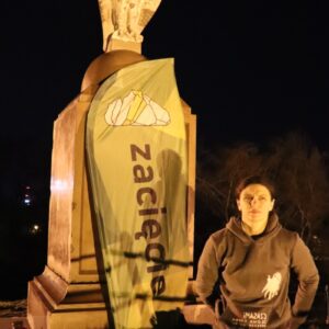 Zajęcie przedstawia kobietę przy pomniku i fladze reklamowej.